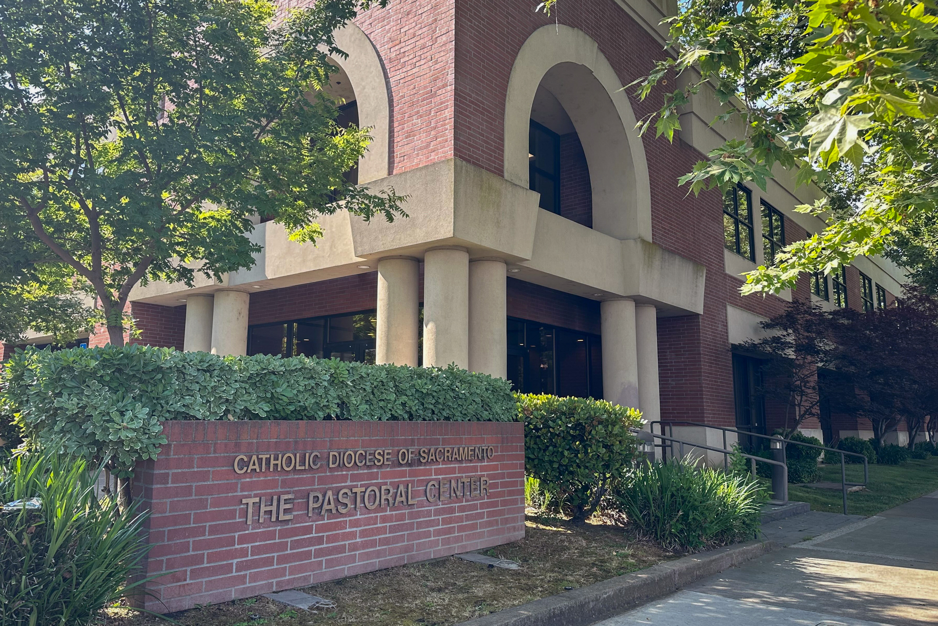 A brick building with verdant landscaping on a sunny day and a brick sign reading "Catholic Diocese of Sacramento: The Pastoral Center."