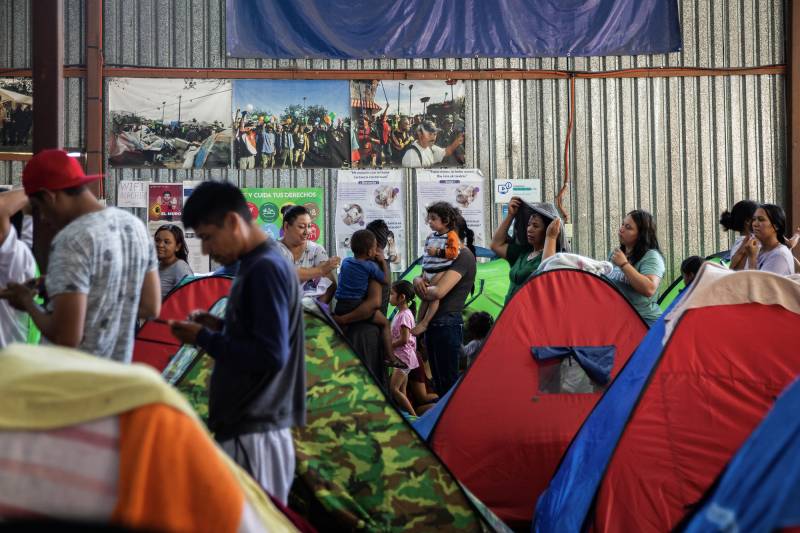 A group of people of various ages standing in line with some holding children near colorful tents in a warehouse.
