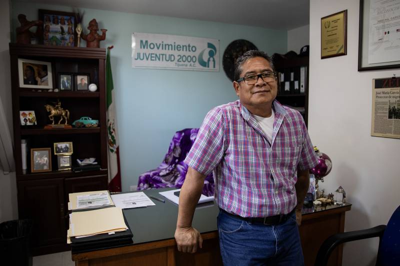 A man wearing glasses and a purple and white checkered button-down shirt and blue jeans leans with both arms against a desk in an office.