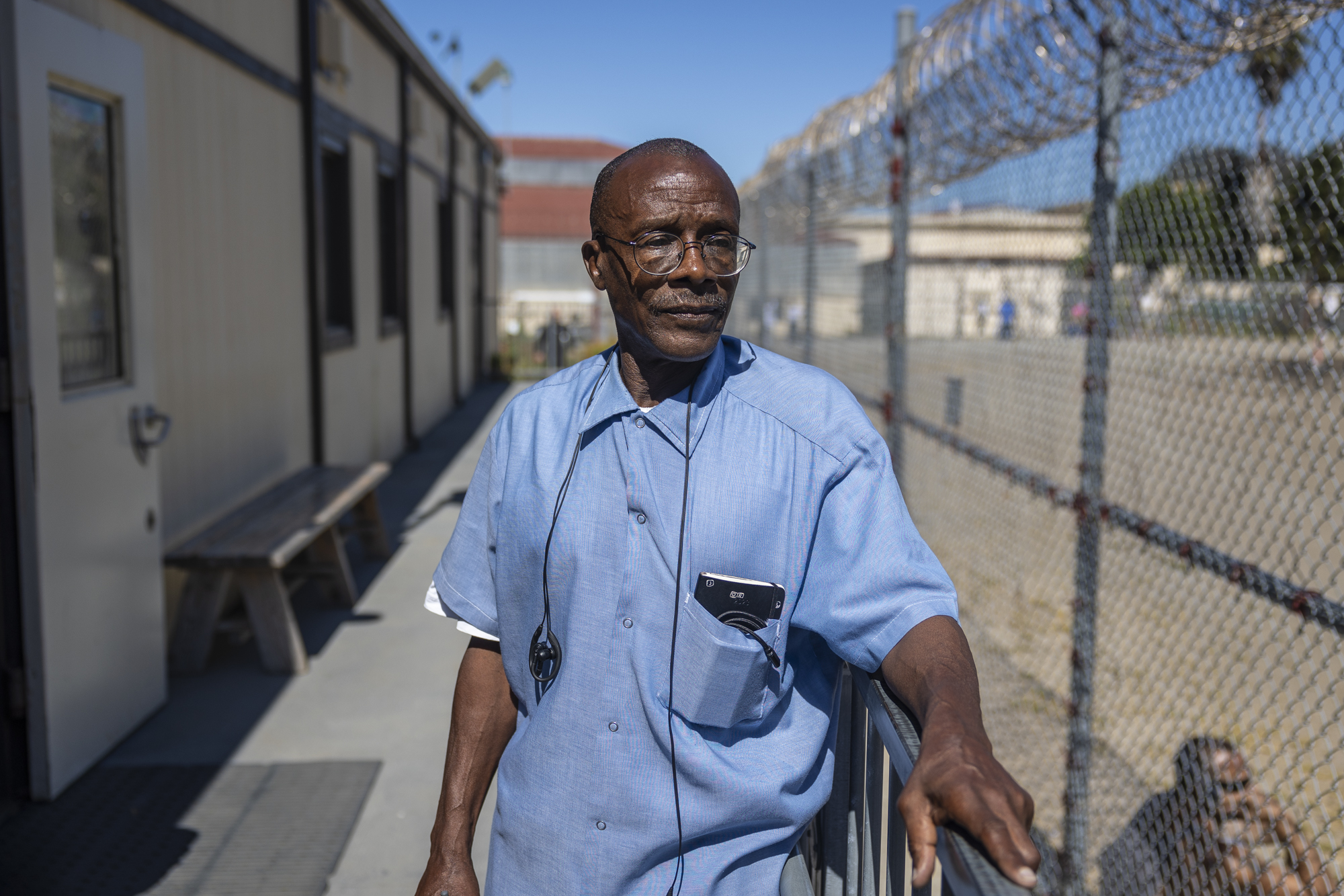 A man with glasses outdoors anA man with glasses outdoors and beside a chain-link fence topped with barbed wire.d beside a chain-link fence topped with barbed wire.