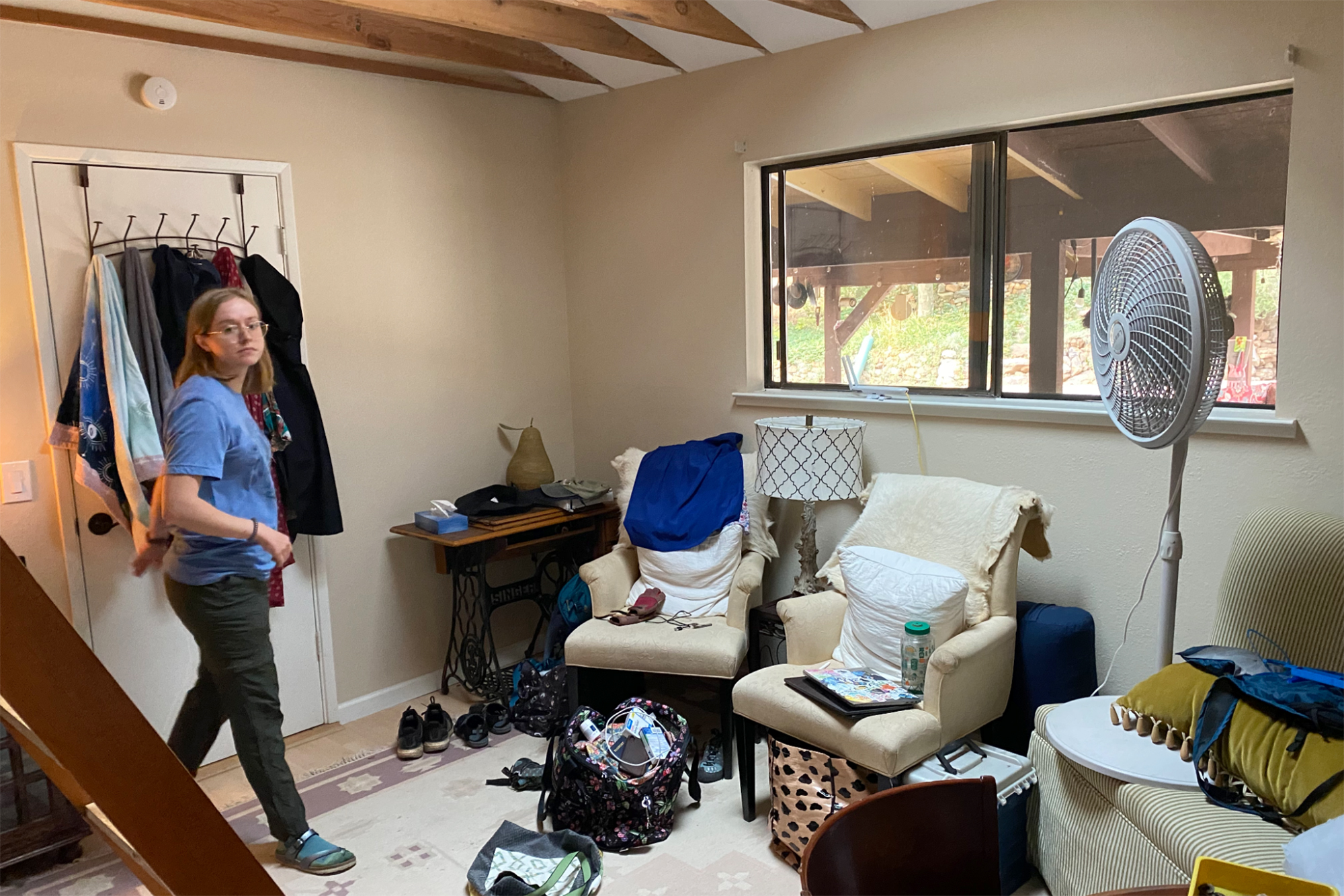 A person with glasses inside a room with clothes and chairs and a tall fan.