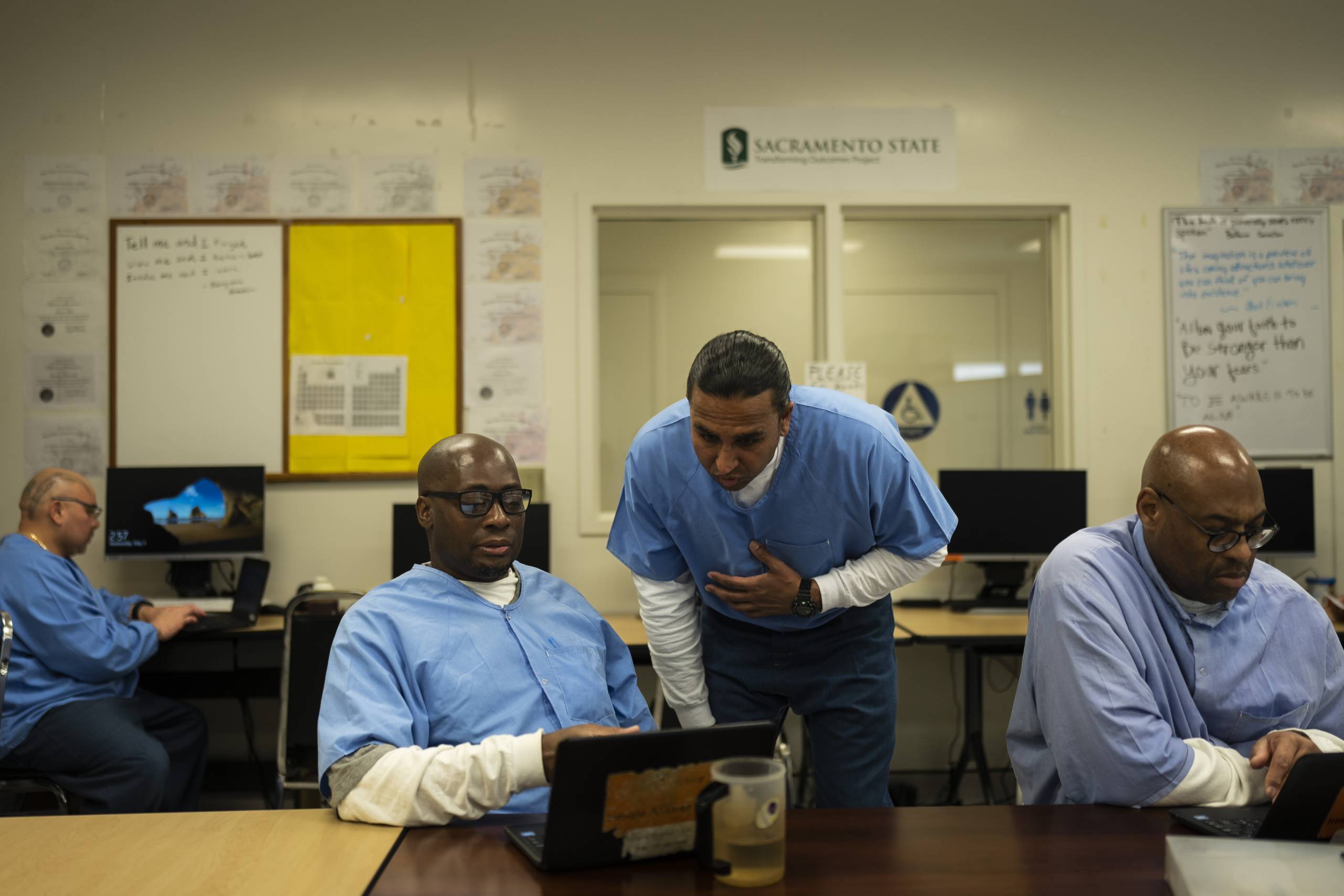 A Black man in a blue prison shirt sits at a table in front of a laptop in a classroom, while another man, of South Asian descent, also in a prison shirt, looks over his shoulder.