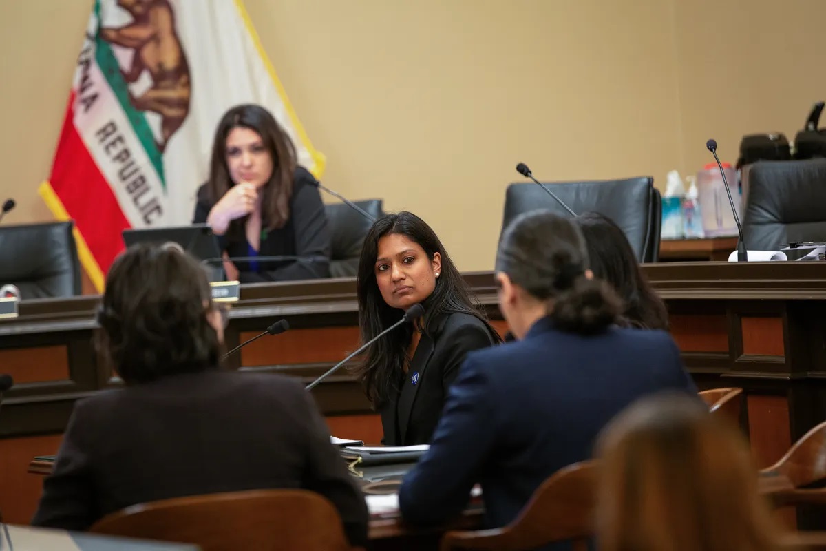 Inside the Assembly Judiciary Committee chambers: There is a California state flag hanging in the background. There are various women in suits and business attire listening to one another. Seats with skinny, black microphones are also seen in the background.