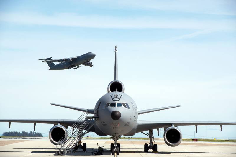 A jet is parked on a tarmac. Another airplane takes off in the sky in the background. It's daytime.