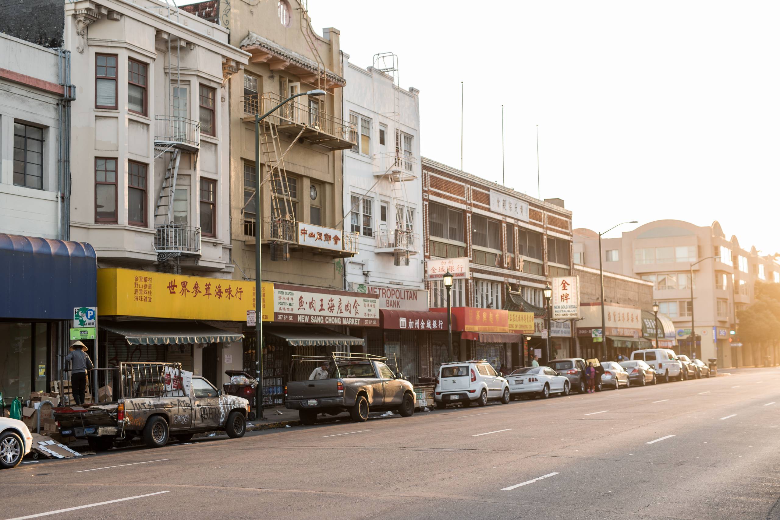 A street in Oakland's Chinatown during the morning. Cars are parked along the street, in front of apartment buildings with shops on the ground level.