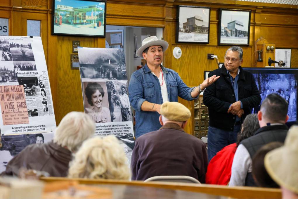 Two men stand in front of a seated audience, showing poster boards with photos on them.