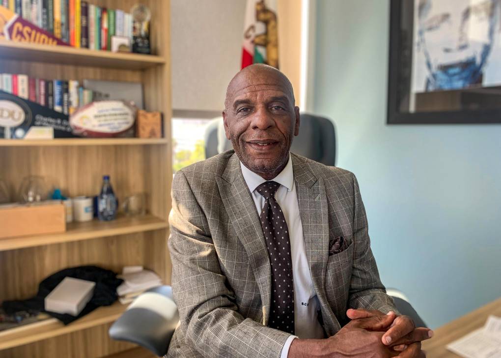 A Black man wearing a jacket and tie, sitting at a desk.