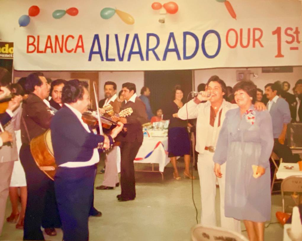 A vintage photo a party with a banner that reads "Blanca Alvarado our 1st" with a woman in a blue dress standing next to a man in a white suit holding a microphone facing a group of men playing instruments.