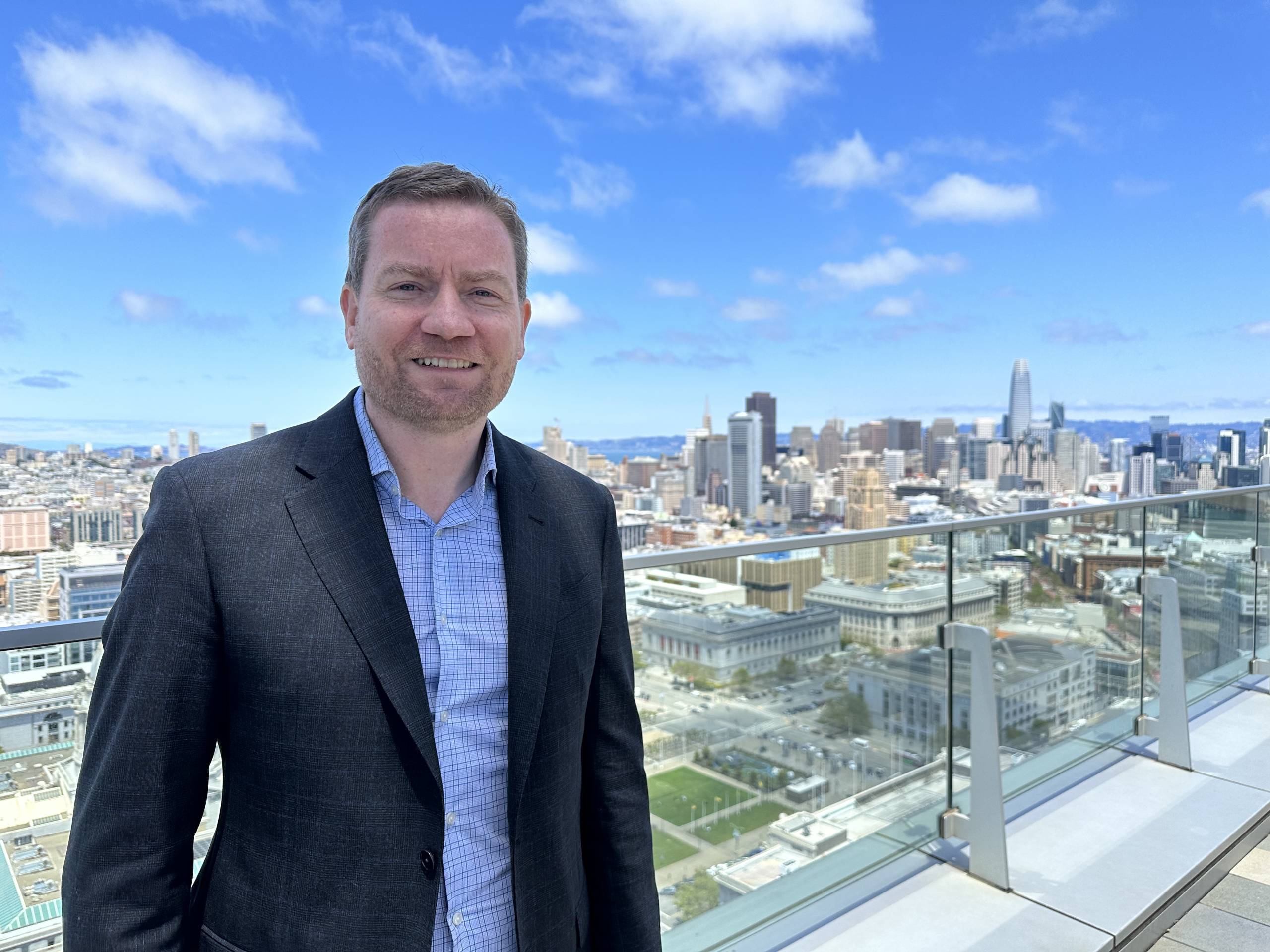A white man wearing a suit smiles at the camera in the foreground. He's standing on the rooftop of a skyscraper. Behind him is a view of the San Francisco city skyline.