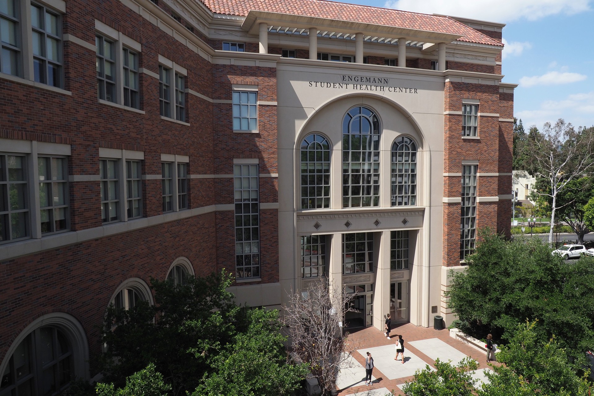 A gigantic, prestigious brick and tan building is pictured on USC's campus. It's the Engemann Student Health Center. Trees surround the front of the building as it hovers over students seen down below entering its glass doors. It's a sunny day with blue skies and puffy clouds.