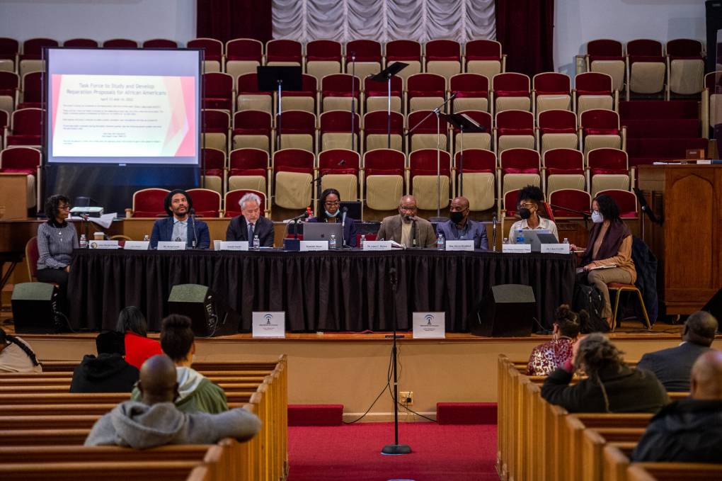 A group of people sit at a long table with microphones in front of them and and a screen to the left behind them with people seated and facing them in aisles.