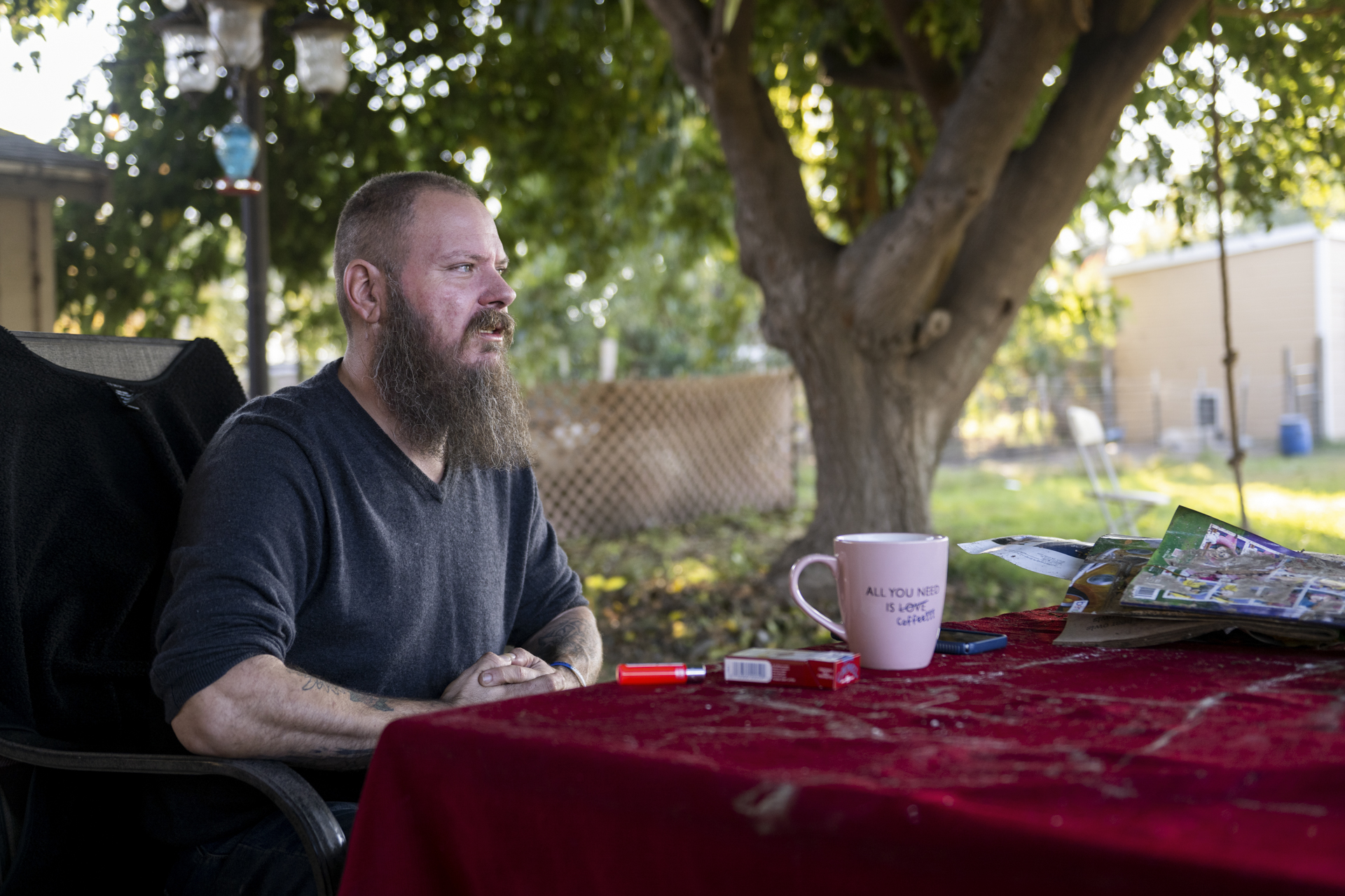 A white man with a long beard sits outdoors in the shade of a tree, at a table with a red table cloth. On the table in front of him are a pack of cigarettes, a lighter, a white mug, a cellphone, and a short stack of papers.