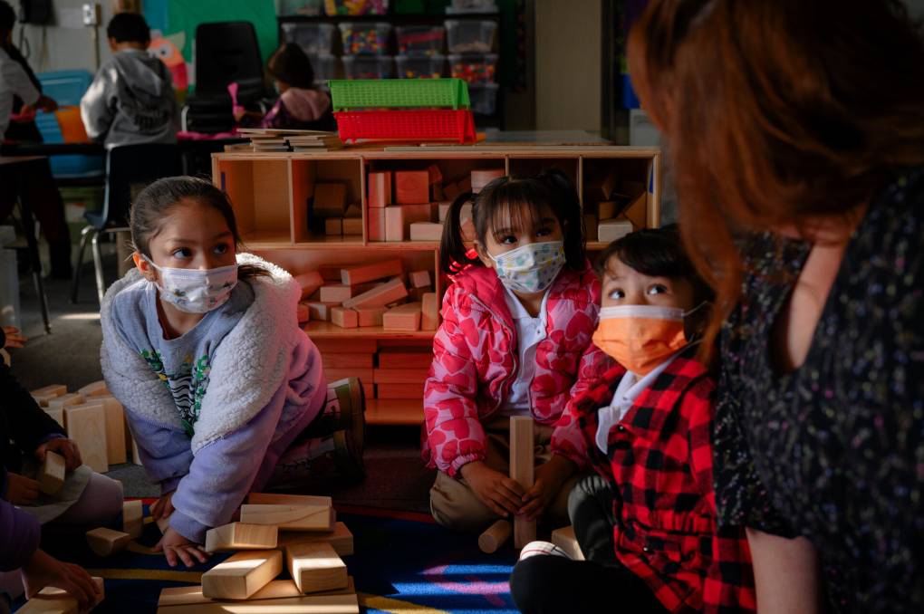 Three kids sit on the floor as they listen to their classroom teacher.