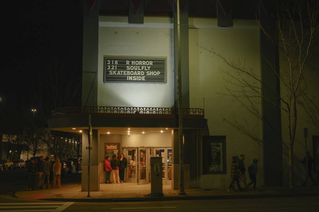 Small groups of people mill about and walk past the exterior of a windowless two-story building with a marquee on it reading "3 18 R HORROR" and "3 21 SOULFLY" and "SKATEBOARD SHOP INSIDE."