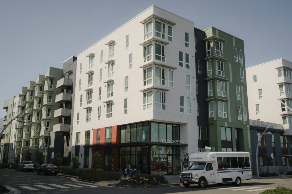 The facade of a multistory green and white residential building seen from across the street. A white shuttle bus is parked in front.