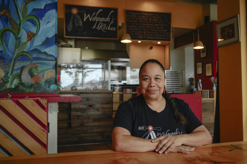 A woman wearing a black t-shirt and with her hair in a braid leans on a wooden countertop and smiles at the camera.