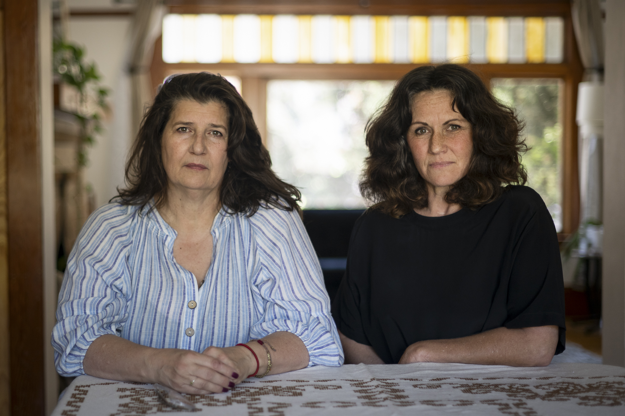 Two people with long hair sit next to each other at a table and look and look at the camera.