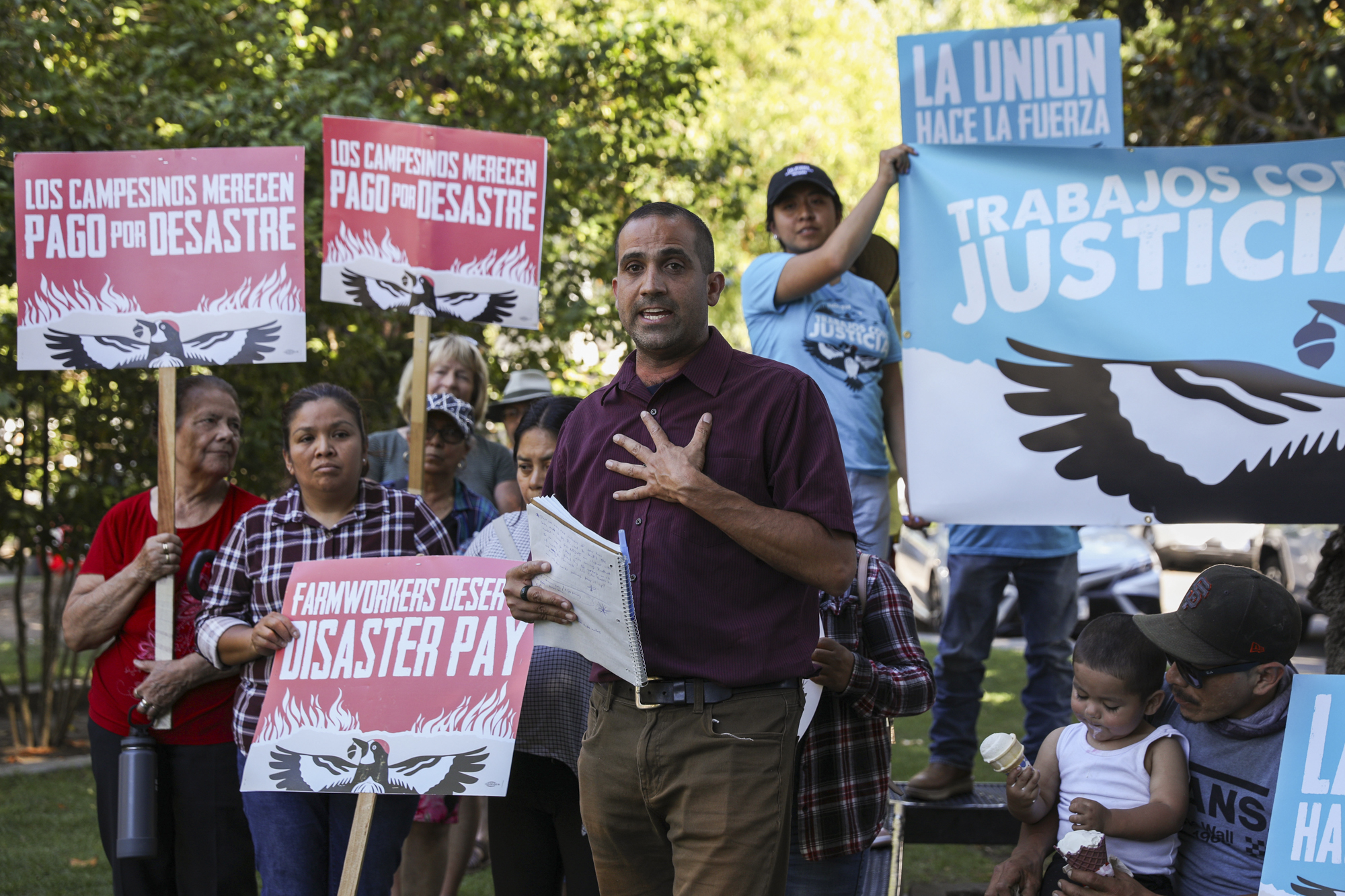 A person with a shaved head speaks in front of others holding signs reading "Farmworkers Deserve Disaster Pay" in an outdoor setting.