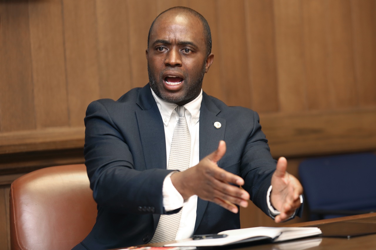 A Black man in a navy business suit, gray tie and white shirt is sitting at a table with a notepad and pen in front of him speaking with his hands extending out. He has a serious face.