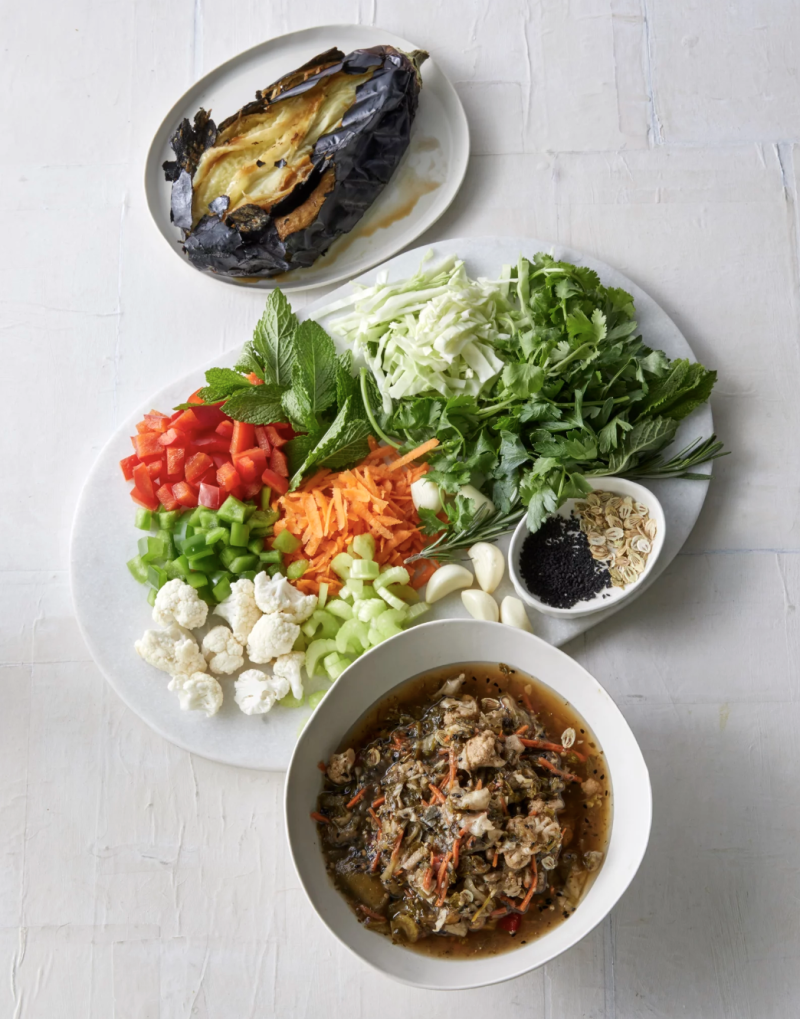 A white plate with chopped vegetables on it sits next to a bowl of stew on a white table.